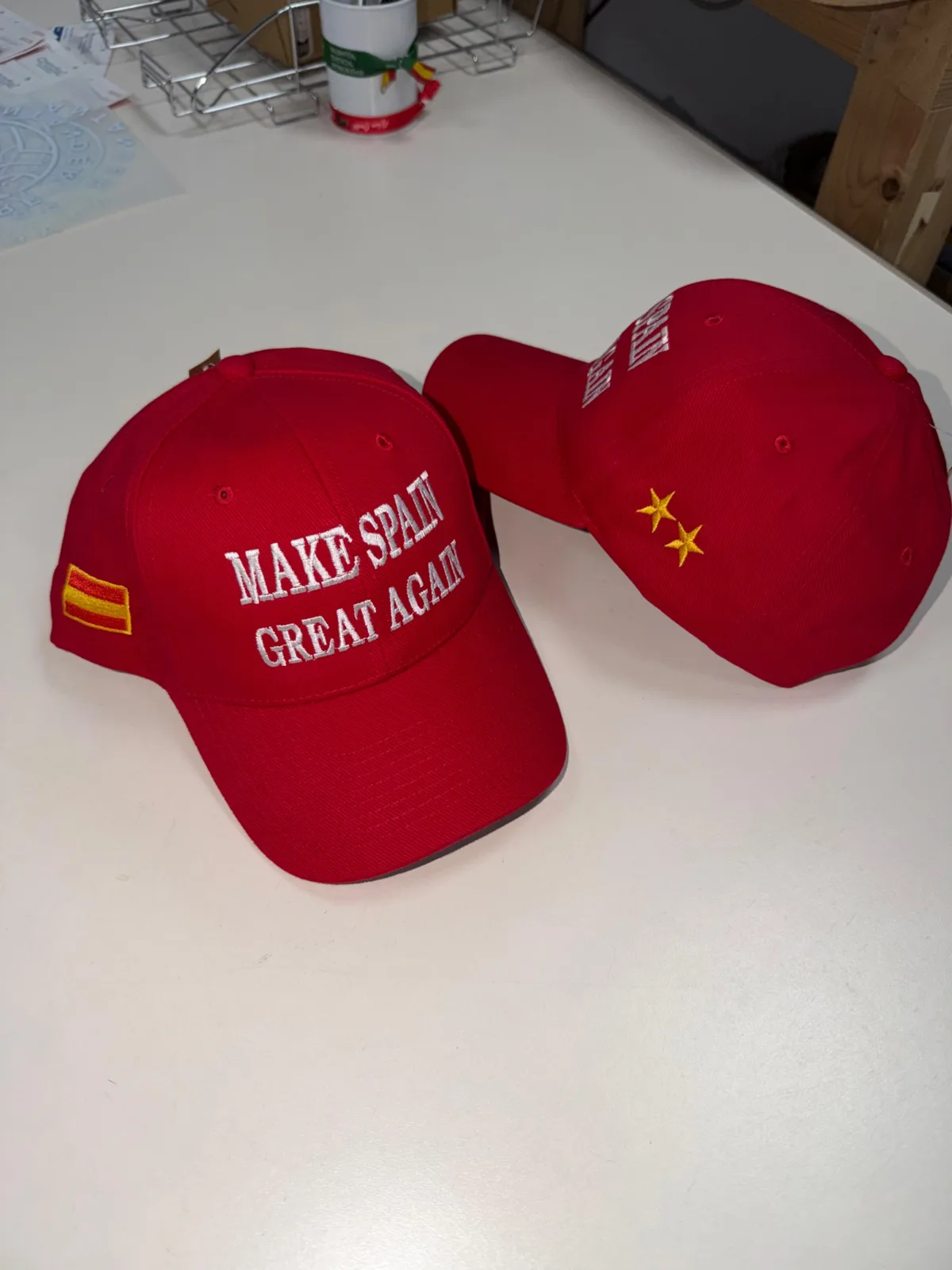 Dos gorras rojas terminadas sobre una mesa, con el bordado frontal, la bandera de España y las estrellas visibles
