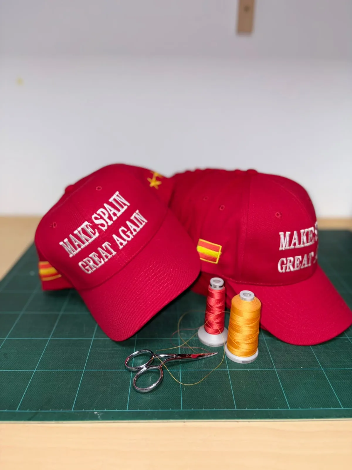 Gorras rojas bordadas en una mesa de trabajo junto a hilos rojo y amarillo y unas tijeras